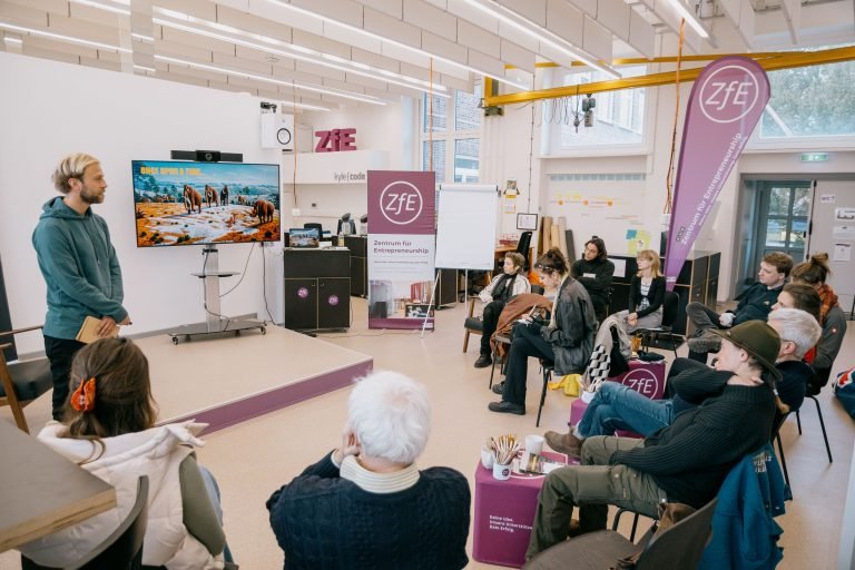 A group of young people sitting in a room listening to a young man speaking to them while standing in the front.