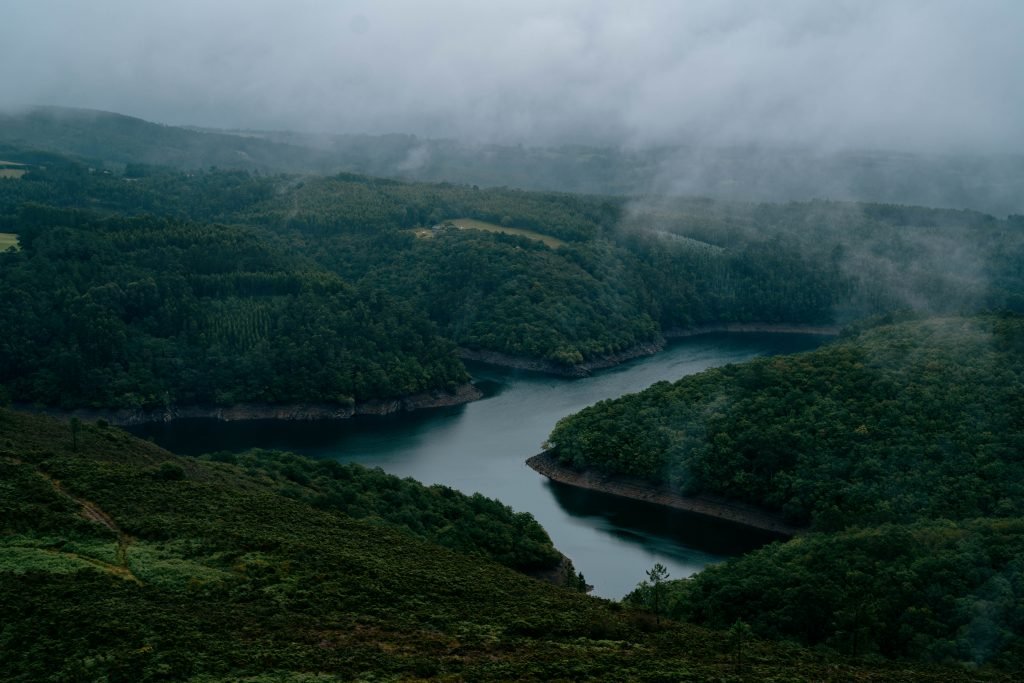 Alternative text: misty landscape featuring a dark blue river running through dark green forested hills